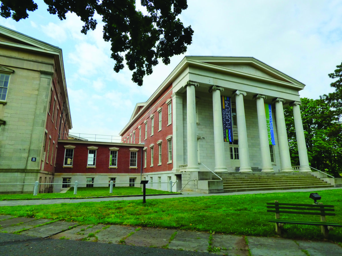 Exterior view of the Staten Island Museum building with greek revival columns and the connecting brick hyphen to the next building. Grass is in the forground
