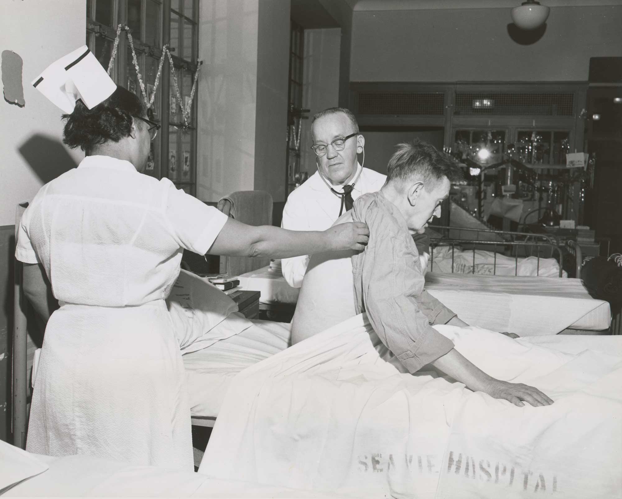 Photo of male patient sitting up as doctor, assisted by nurse, examines him