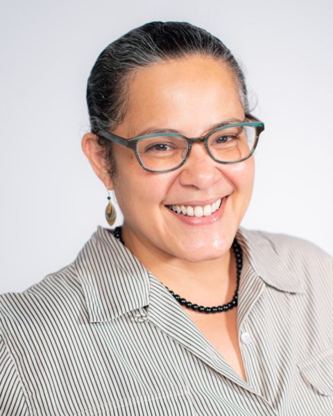 Headshot of woman with black hair pulled back, glasses, grey shirt, smiling