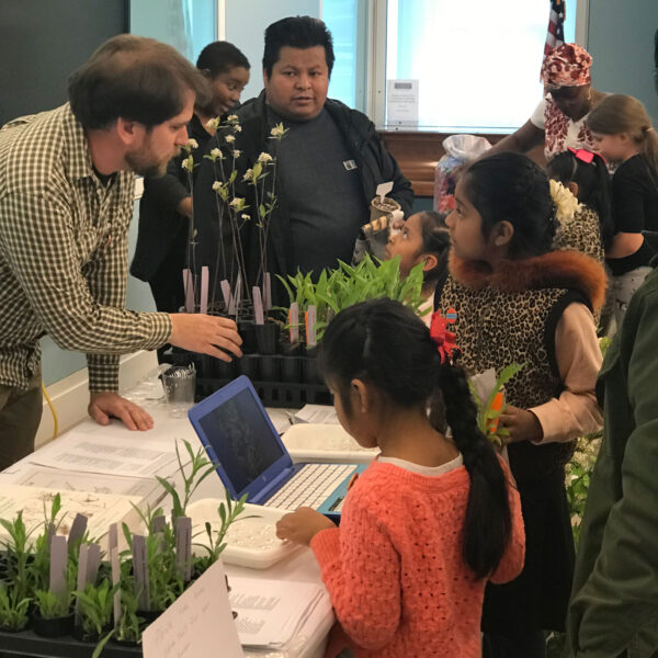 School aged children looking at plants and a lap top on a table while a teacher talks to them