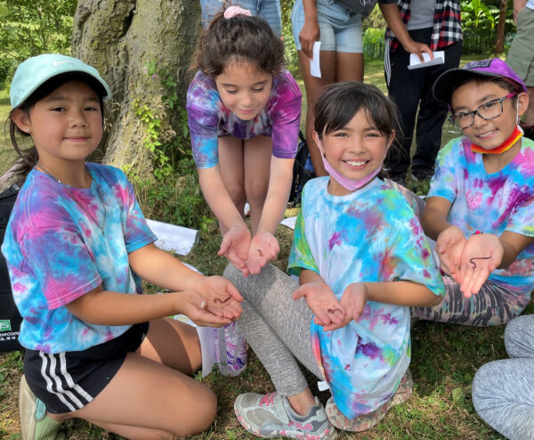 Four school aged girl wearing tie-dyed shirts looking at the camera holding worms in their hands.