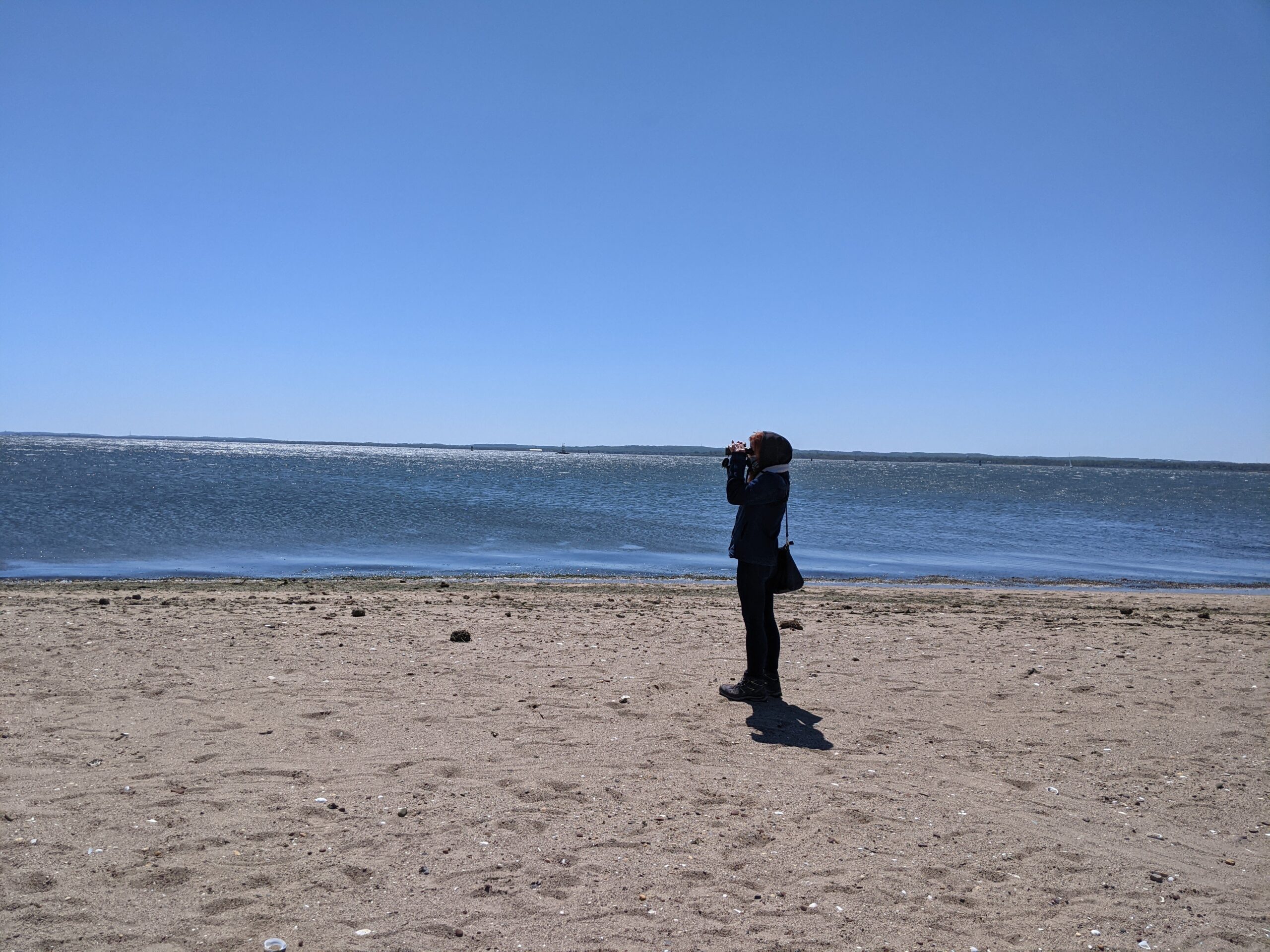 A person standing on a beach looking through binoculars with the water and blue sky behind them