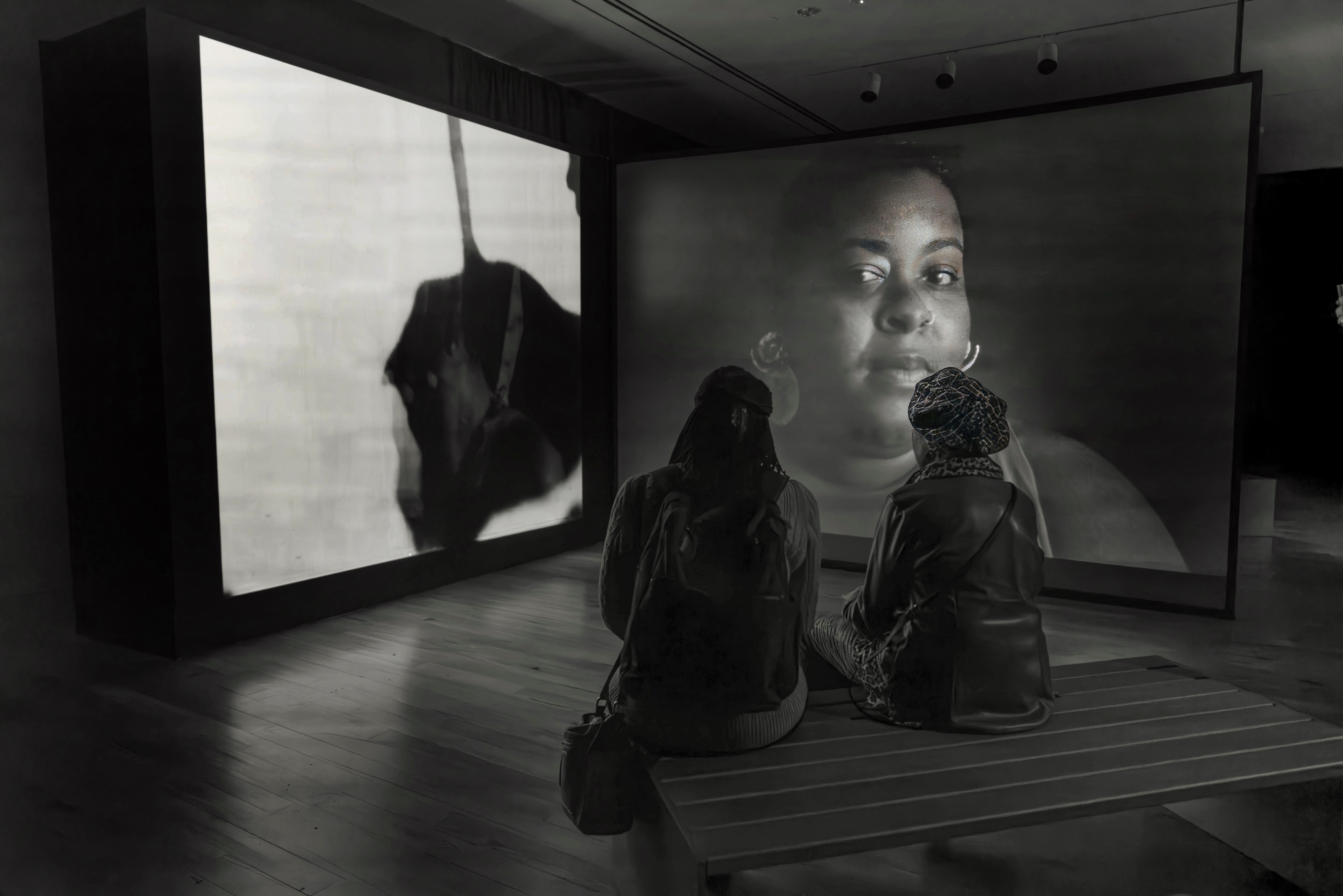Black and white photo of two people sitting on a bench in a gallery watching a four channel film